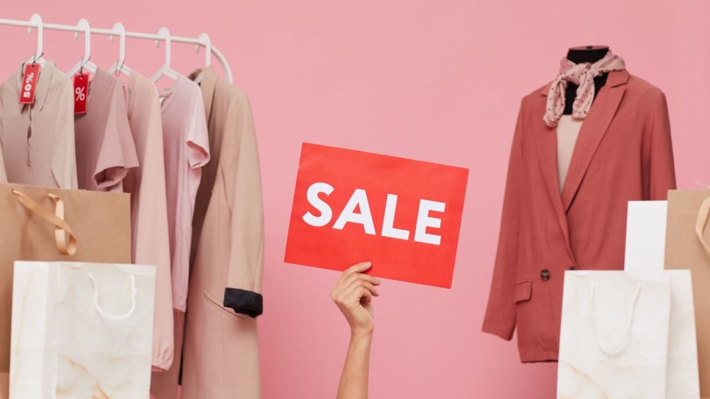 Pink background with womans clothing on racks and an arm in the center holding a Sale sign up