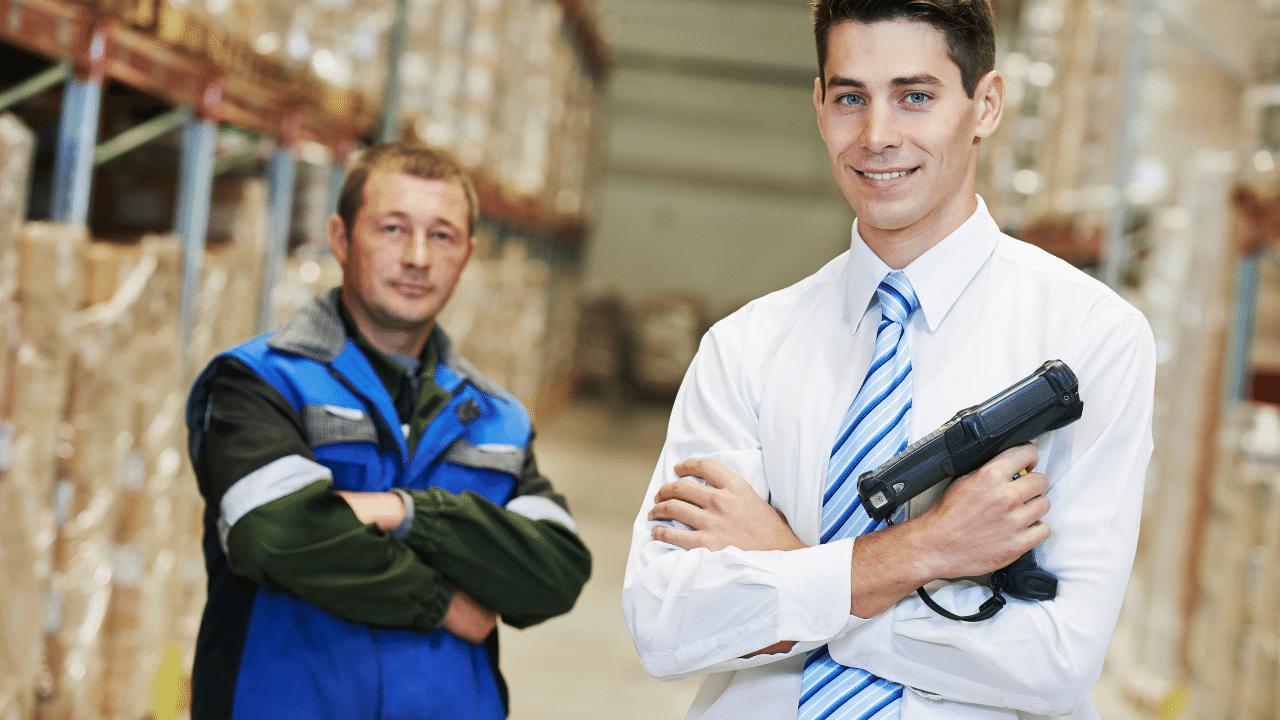 Two men standing in an inventory warehouse with arms crossed, one man in a safety vest, the other man in a tie and dress shirt holding a barcode scanner