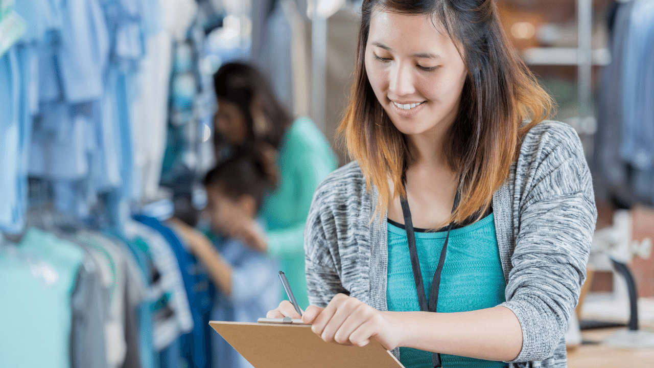 Woman smiling down at a clipboard as she records data while people shop in apparel store in background