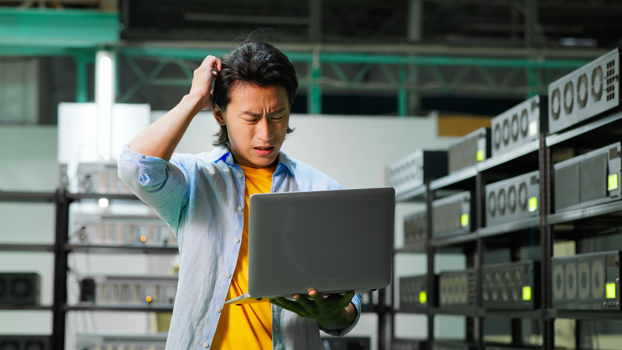 Confused man scratching his head with one hand while holding a laptop in the other while standing in a crowded server room